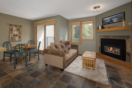 A cozy living room with a brown sofa, a small wooden table, and a fireplace. There's a dining area with four chairs and a table. Large windows let in natural light and offer a view outside.