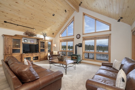 A spacious living room with wooden vaulted ceilings, large windows, leather sofas, a wooden entertainment center, and a fireplace.