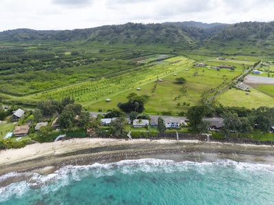 Aerial view showcasing beachfront properties nestled between pristine turquoise waters and lush tropical landscapes with rolling hills and agricultural fields.