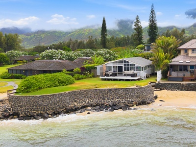 A stunning beachfront property nestled between emerald mountains and crystal-clear waters, with lush tropical gardens and a spacious lawn leading directly to a private sandy shore.