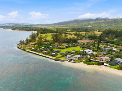 Aerial view showcasing a serene coastal neighborhood with lush green lawns, sandy beach access, and mountain vistas rising in the background under a clear blue sky.