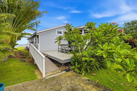 Coastal cottage featuring covered deck seating area, surrounded by tropical palms and flowering plumeria, with blue skies overhead and open lawn leading to waterfront.