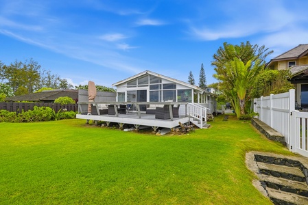 A modern beach house featuring an expansive wooden deck with comfortable outdoor seating, surrounded by lush tropical landscaping and mature palm trees under clear blue skies.