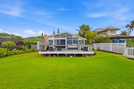 Charming beachfront cottage features a spacious outdoor deck and lush green lawn, surrounded by swaying palms and tropical landscaping in a peaceful residential area.