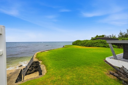 The property features a pristine oceanfront lawn stretching to a private sandy beach, with calm blue waters meeting the horizon under clear skies.