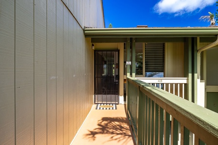 The property entrance is marked with unit number 48, featuring a welcoming covered porch and green railing beneath a bright blue sky and palm trees.