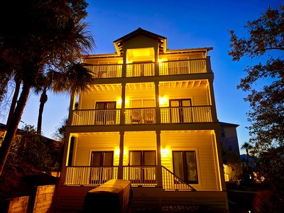 Your coastal retreat glows warmly against twilight sky, surrounded by tropical palms and mature trees in peaceful neighborhood.