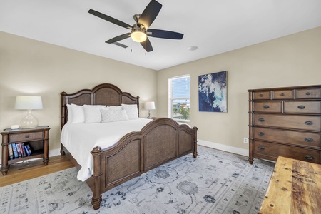 Your peaceful retreat awaits with crisp white linens, warm wood furnishings, and a ceiling fan keeping you comfortable. Natural light filters through the window while coastal-inspired decor adds the perfect finishing touch.