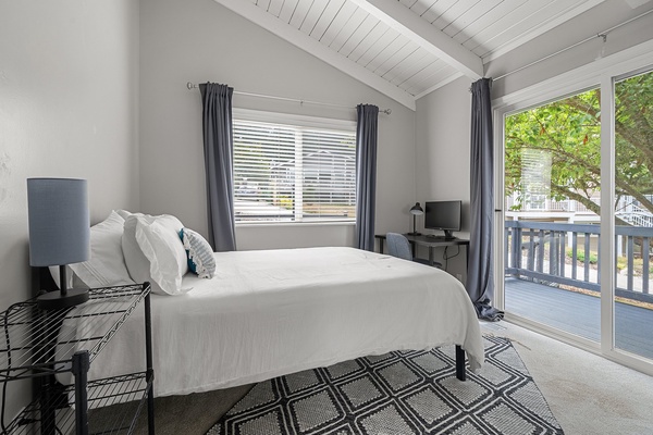 Cozy third guest bedroom with vaulted ceilings, and a small workspace next to a private balcony.