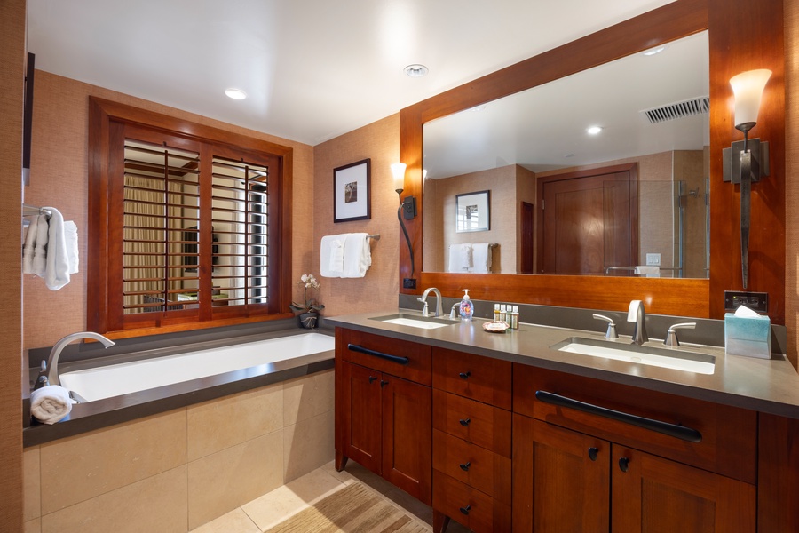 Unwind in your luxurious bathroom featuring warm wood tones, dual vanities, and a deep soaking tub by elegant shuttered windows.