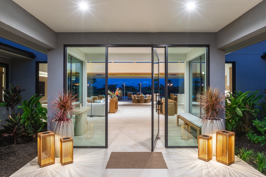 Step through elegant glass doors into your luxurious retreat, where warm lighting and tropical plants create an inviting welcome.