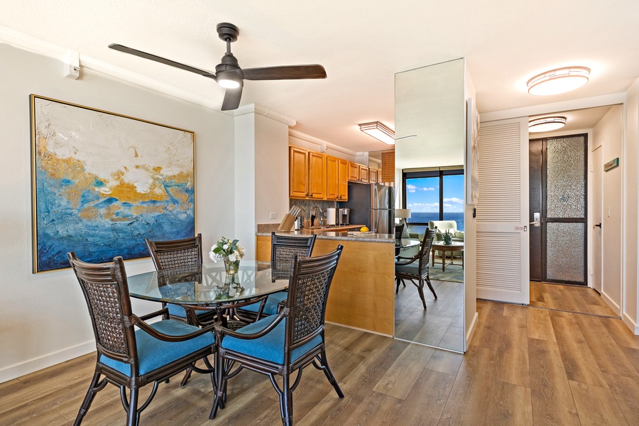 The dining area features a glass-top table with seating for four, complemented by a modern ceiling fan and a vibrant piece of artwork.