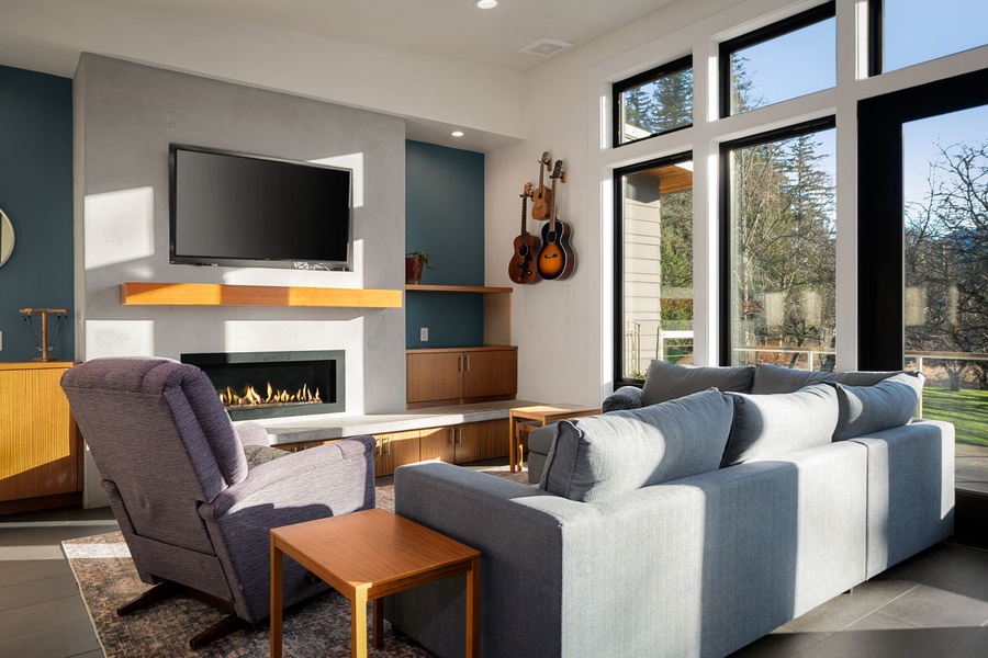 Relax in your modern living room with cozy seating, a crackling fireplace, and expansive windows framing beautiful garden views.