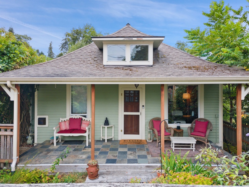 Charming cottage with welcoming covered porch featuring comfortable seating and colorful cushions surrounded by lush greenery.