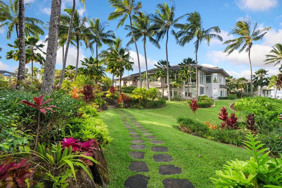 A winding stone path leads you through a meticulously manicured garden, adorned with vibrant blooms and towering palm trees.