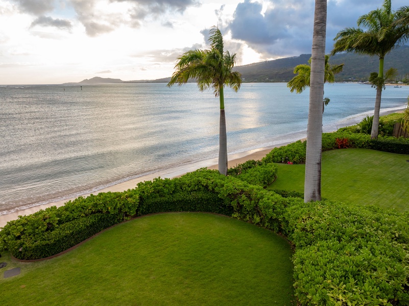 Beachfront property featuring manicured lawns bordered by tropical palms, with pristine sands and calm turquoise waters stretching toward distant mountains under dramatic coastal skies.