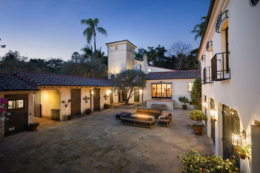 Spanish-style villa features an elegant courtyard with fire pit seating and terracotta tile roofing under twilight skies.