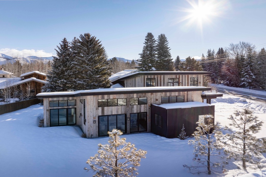 Your winter cabin surrounded by snow-covered pines, with the sun peering over the rooftop.