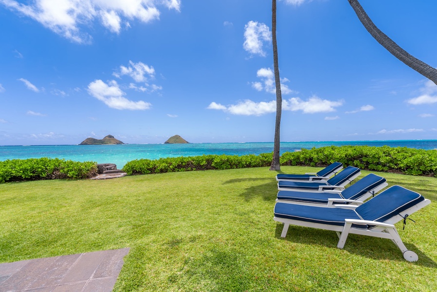 A pristine beachfront property in a tropical coastal setting with lush lawn, turquoise ocean waters, small offshore islands, and palm trees framing the view under blue skies.