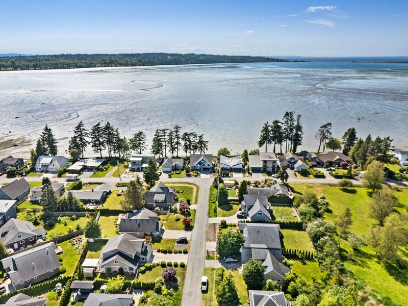 Aerial view of a waterfront neighborhood with homes nestled among tall evergreens, overlooking a serene bay and distant forested shoreline under clear blue skies.