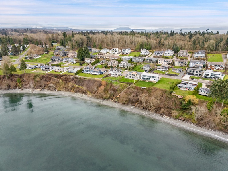 Aerial view of waterfront residential community with homes along the shoreline, surrounded by forests and distant mountains.