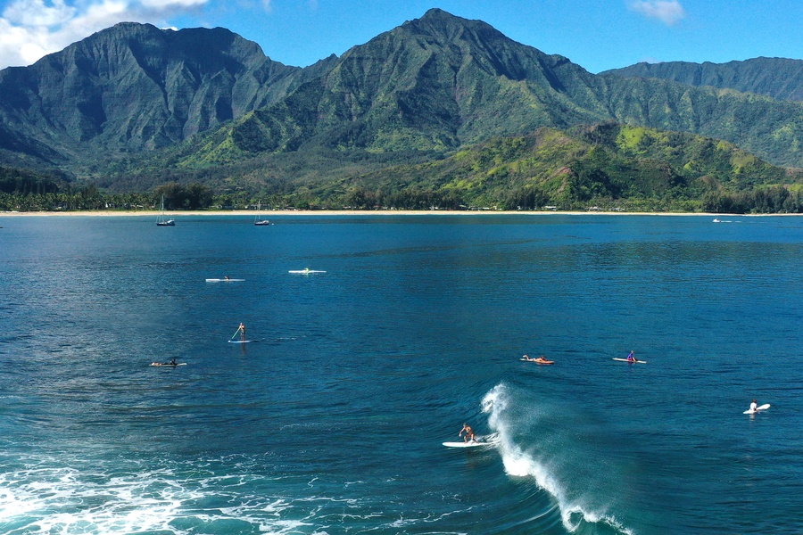 Surfers catching waves at Hanalei Bay with dramatic mountain backdrops and crystal-clear waters.