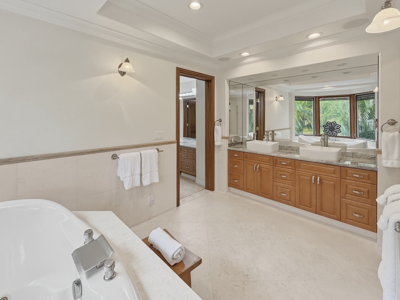 Spa-style bathroom with soaking tub, dual vanities, and tropical garden views through warm wood-framed windows.