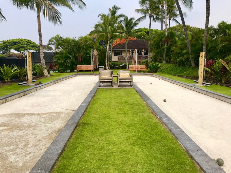 The Hualalai Resort bocce ball court featuring manicured grass lanes, palm trees, and comfortable seating in a lush garden setting.