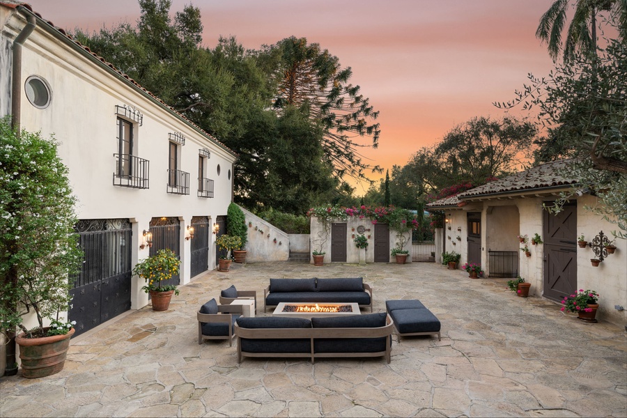 Charming Spanish-style courtyard with fire pit seating, surrounded by white adobe buildings and lush gardens under a golden sunset sky.