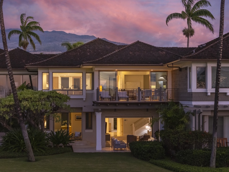 Tropical villa at dusk with mountain backdrop, palm trees, and warm interior lighting creating an inviting Hawaiian atmosphere.