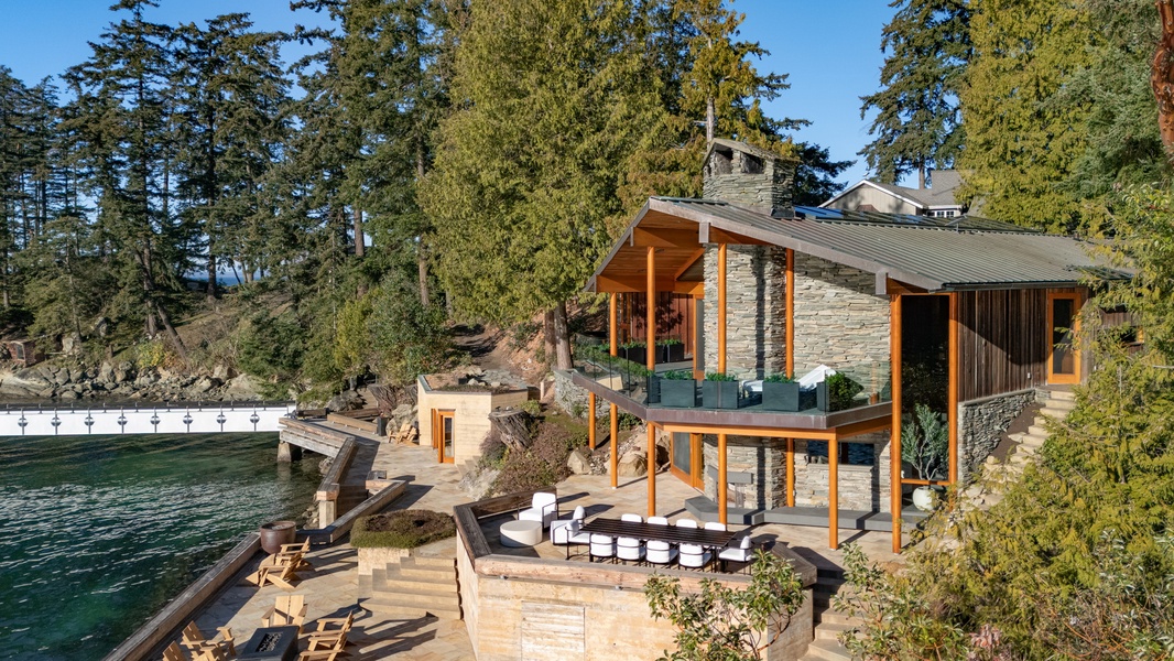 Modern waterfront property nestled among towering evergreens, featuring natural stone and warm wood accents with multiple outdoor terraces overlooking pristine waters.