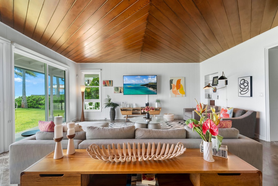 Relax in your spacious living room where vaulted wood ceilings and tropical light create the perfect retreat. Floor-to-ceiling glass invites lush greenery and ocean breezes inside, while vibrant art and plush seating surround you in coastal comfort.