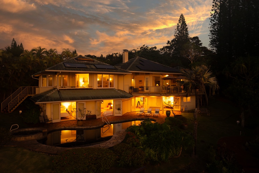 Stunning tropical villa glows warmly beneath dramatic sunset skies, surrounded by lush vegetation and mature palm trees.