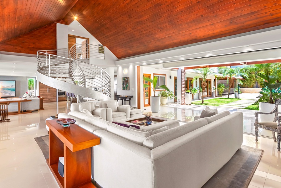 Relax in your spacious living room beneath stunning wood-paneled ceilings, where modern comfort meets tropical elegance and lush garden views await just beyond the open doors.
