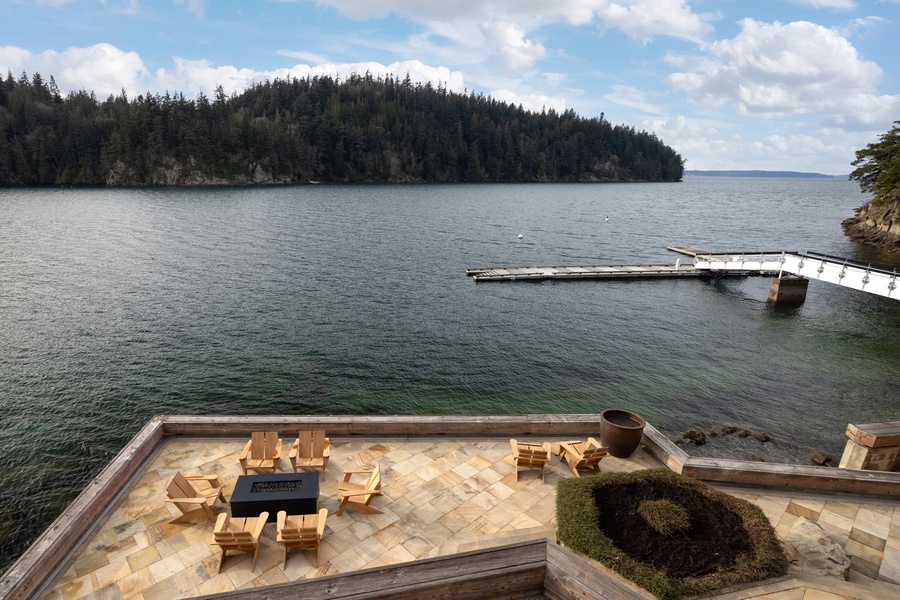 Expansive lakefront terrace features natural stone flooring, wooden outdoor furniture, and panoramic water views with forested islands stretching across the horizon.