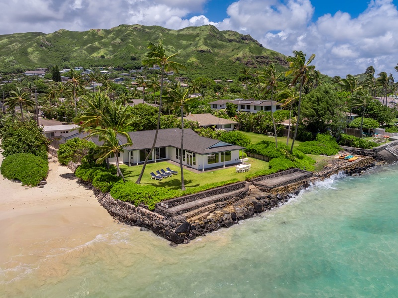 Stunning beachfront property nestled between turquoise waters and lush green mountains, offering direct beach access and tropical landscaping with swaying palms and sunbeds in a serene Hawaiian coastal neighborhood.