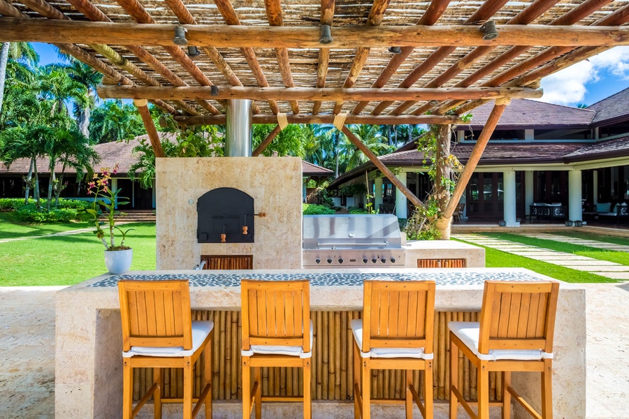 Outdoor kitchen area with a stone oven and stainless steel grill, featuring a wooden bar stools for casual dining.