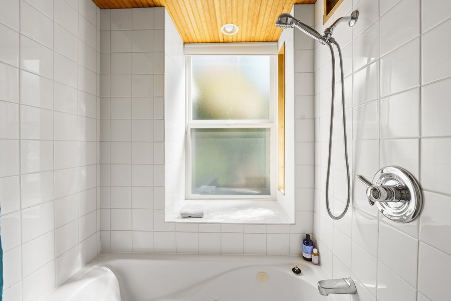 Unwind in your bright, tiled bathroom featuring a deep soaking tub and handheld shower beneath warm wood ceiling accents.