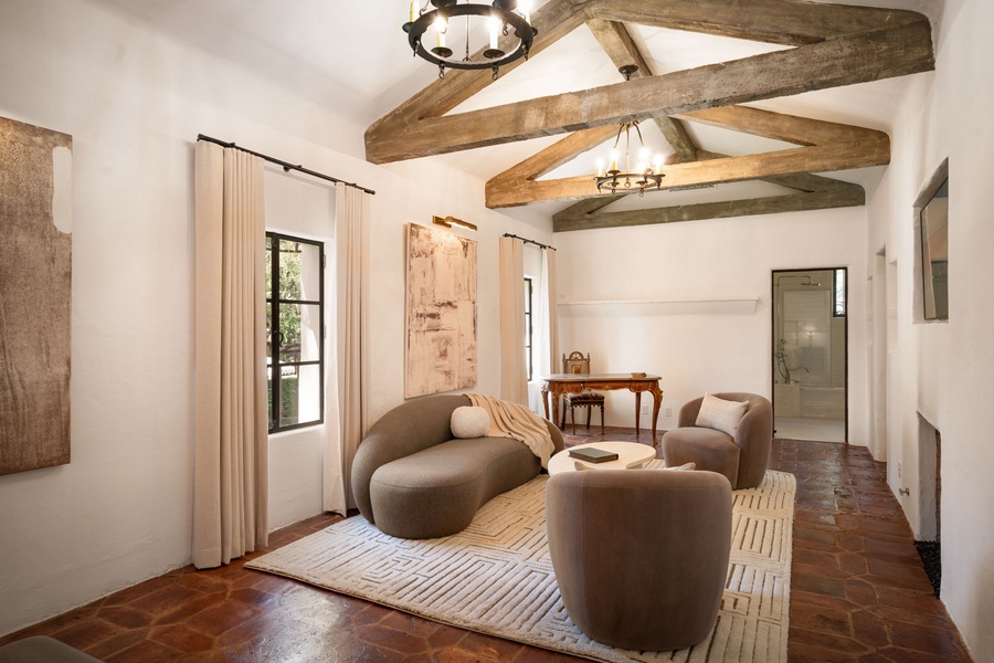 Your elegant lounge area in the primary suite features exposed wooden beams, plush curved seating, and natural light streaming through French doors.