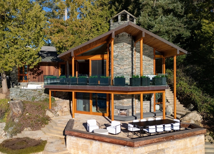 Modern lakeside retreat featuring stone and wood architecture with multiple outdoor terraces surrounded by mature trees and natural landscaping.