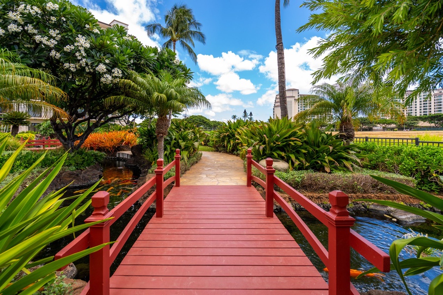 Tropical garden oasis with charming red bridge spanning peaceful waters, surrounded by lush palms and vibrant blooms.