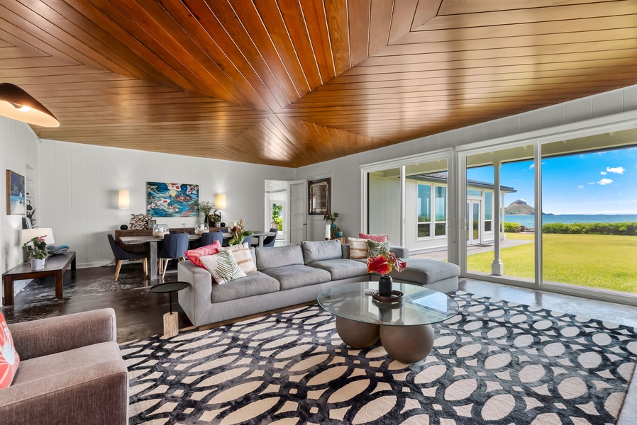 Step into your expansive open-plan living space, where soaring wood ceilings and floor-to-ceiling windows frame stunning ocean and garden views. Unwind on the plush sofa or gather at the dining table—relaxation awaits in every corner.