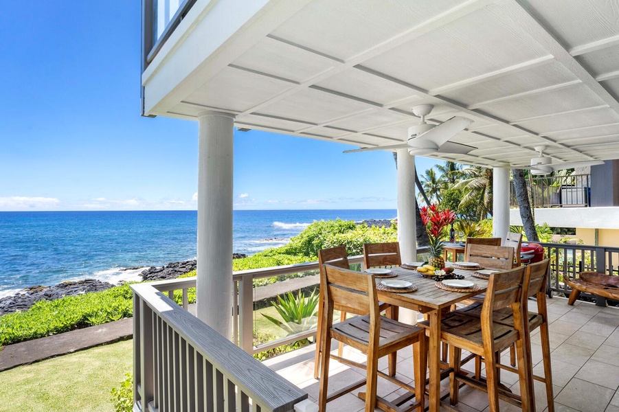 Lanai dining area framed by tropical foliage, beach stone, and ocean horizon.
