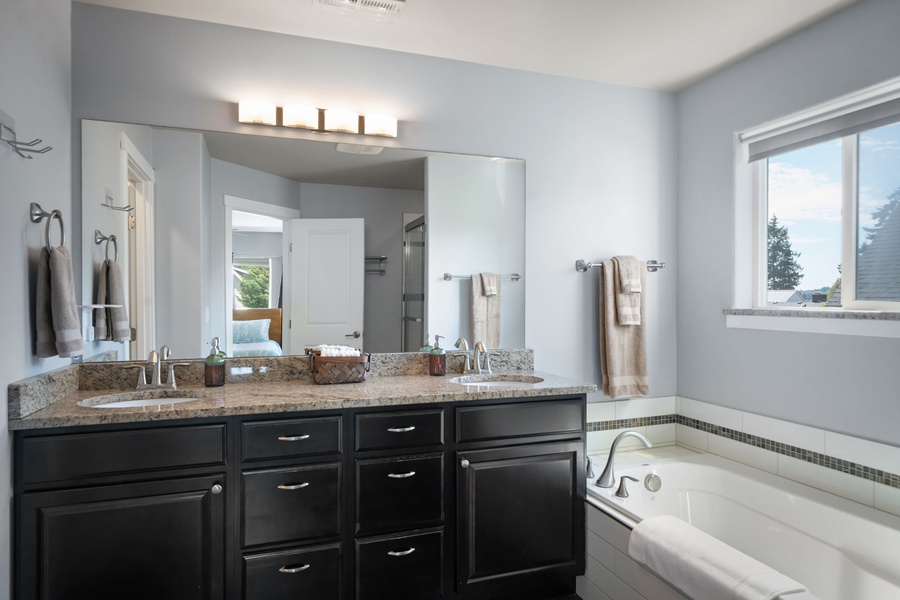 Your private bathroom features a luxurious double vanity with gleaming granite countertops, a soaking tub perfect for unwinding, and soft natural light streaming through the window. Everything is ready for your relaxing getaway.