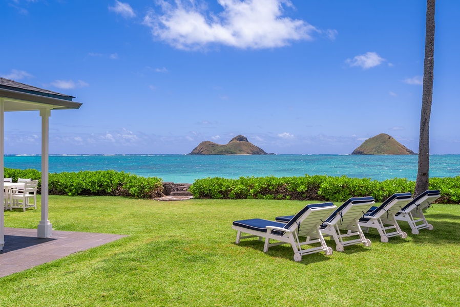Beachfront lawn with loungers offers stunning views of turquoise waters and offshore islands, framed by lush tropical landscaping and partial shade structure.