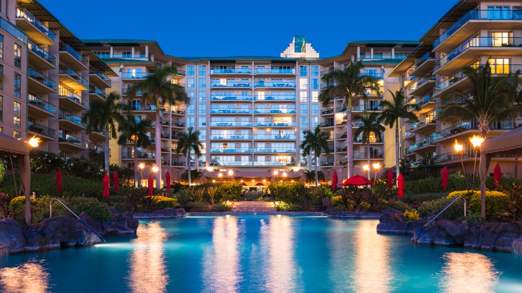 This stunning view showcases a beautifully illuminated resort at dusk, with the pool in the foreground reflecting the lights and surrounding palm trees