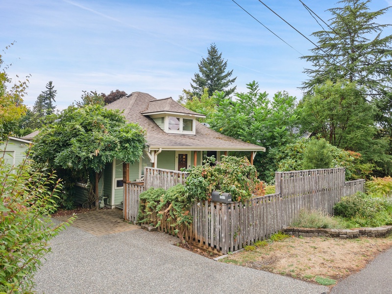 Charming green cottage nestled among mature trees with private fenced yard and covered front porch in peaceful residential neighborhood.