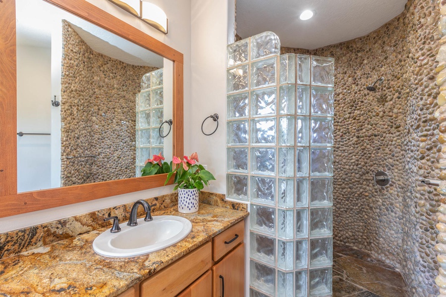 Stone-accent ensuite bath at Pohaku Villa with glass block shower and rustic vanity design.