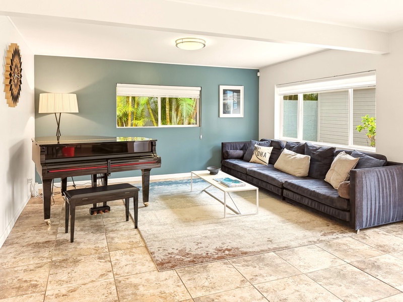 The spacious living room features a plush sectional sofa, elegant piano, and tropical palm views through large windows.