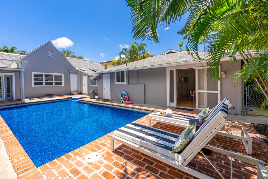 A secluded outdoor retreat featuring a sparkling private pool, sun-soaked brick deck, and shaded loungers framed by tropical palms—your perfect spot to unwind under blue skies.